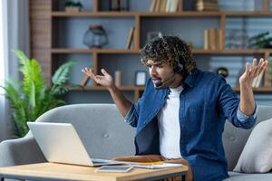 Dissatisfied man sitting on sofa at home, with headset and headphones. Nervous freelancer working remotely using call, arguing and shouting. photo