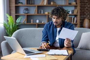 Man working at home with documents, bills and checks. Using laptop and calculator to calculate utility bills, rent. Sitting on sofa in living room of apartment. photo
