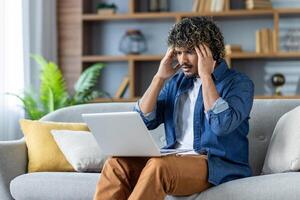 Upset man sitting on sofa in living room at home, in apartment. Employee working remotely using laptop, received notification, error message. photo