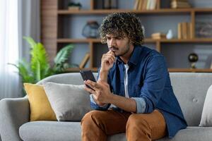Serious thinking man with a phone in his hands sits on a sofa in the living room of an apartment. He reads messages and browses social networks thoughtfully and concentratedly, uses an application. photo