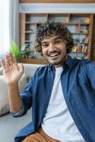 Vertical shot, man smiling and looking at camera, waving hello, using app on phone for call. Sitting at home in living room of apartment. photo