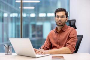 Confident businessman working at laptop in modern office. Man in casual attire focused on tasks. Bright, professional workspace with large windows. Concept of technology, business, and productivity. photo