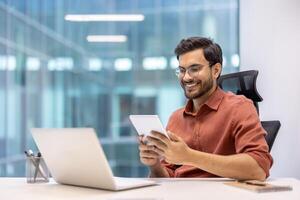 Professional sitting at office desk using tablet while working on laptop. Modern corporate environment with large windows. Concept of productivity, technology, and business success. photo