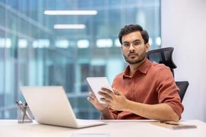 Serious programmer holding tablet in modern office setting, working on digital project with laptop. Business professional in smart casual attire, concentrating on work tasks. photo