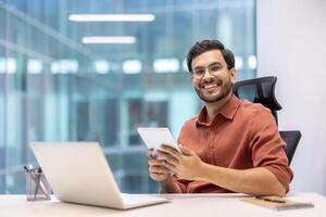 Smiling young professional man sitting at desk using tablet and laptop in modern office environment. Workspace includes notebook and phone. Concept of technology, business, productivity photo