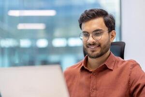 Smiling professional man using laptop in modern office. Confident individual in glasses wearing red shirt while focusing on work tasks. Represents productivity and workplace satisfaction. photo