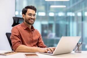 Smiling man wearing glasses and an orange shirt working on laptop in modern office. Phone and notebook on desk. Bright and professional workspace with glass walls. photo