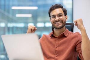 Happy young professional celebrating success at desk in modern office setting. Showing excitement and achievement while working on laptop. Positive work environment and career growth concept. photo