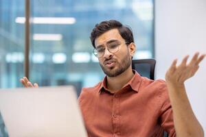 Man sitting at desk using laptop in office, looking frustrated and confused. Feeling of stress and uncertainty. Business-related emotions. photo