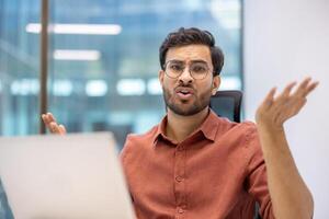 Confused man working on laptop in modern office. Expressing uncertainty and frustration. Professional atmosphere with glass windows and natural light. Wearing glasses, using phone and computer photo