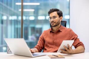 Serious businessman working with laptop and tablet in modern office. Focused on tasks, using both devices to enhance productivity. Professional setting with large windows and natural light. photo