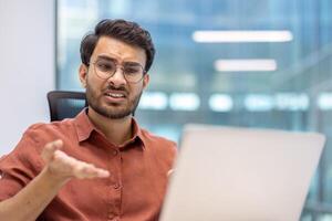 Young man experiencing frustration while working on laptop in modern office setting. Male professional gesturing with hand, indicating confusion or displeasure during work. photo