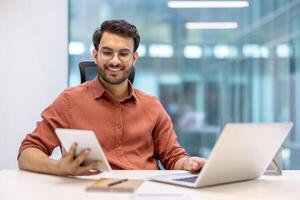 Smiling young man in office setting working with tablet and laptop. Displays confidence and modern workplace concept, illustrating multitasking and productivity. Image ideal for business, technology photo