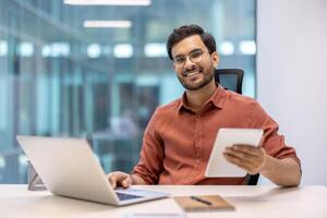 Indian programmer working in modern office with laptop and tablet. Professional smiling, using technology for tasks, productivity. Contemporary workspace, glass walls, tech gadgets. photo