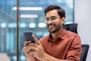 Cheerful man wearing glasses holding and using smartphone in office environment. Happy and relaxed expression while interacting with phone. Professional attire with orange shirt photo