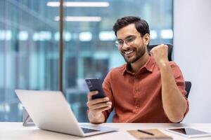 Excited businessman celebrating success at work using smartphone. Young professional sitting at office desk with laptop, phone, and notebook, showing joyful expression and happiness. photo