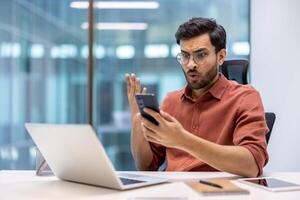 Man sitting in modern office, looking surprised at phone message while working on laptop. Expression shows shock and disbelief. Office environment with digital devices and workspace. photo