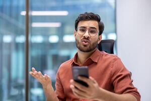 Office worker expressing frustration during phone call in modern office. Professional being annoyed. Business person handling important call in workplace environment. photo