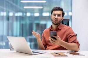 Man in office showing confusion holding smartphone, expressing frustration during work. He is sitting at desk with laptop and tablet, conveying business communication or technology issues. photo