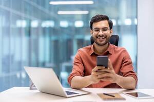 Smiling man sitting at office desk using smartphone for work communication in modern corporate environment. Person enjoying remote work convenience with laptop and phone technology. photo