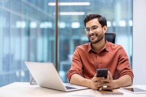 Businessman smiling while working on laptop and smartphone in modern office setting. Confident professional multitasking at bright workplace. Glass walls and contemporary design in background. photo