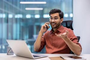 Confident man in office setting engaged in phone call while using laptop. Displays elements of technology, communication, business professionalism. Captures essence of modern work environment photo