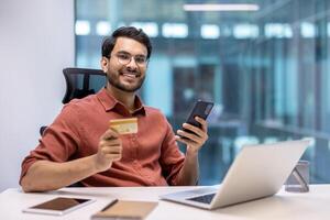 Smiling man in office holding phone and credit card at desk with laptop. Concept of online banking, digital payment, work, and technology. Confident and happy professional connecting digitally. photo