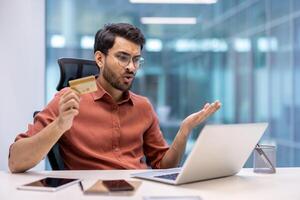 Man sitting at desk holding credit card while using laptop, showing frustration and confusion with online transaction. Office environment suggests business or personal finance issues. photo