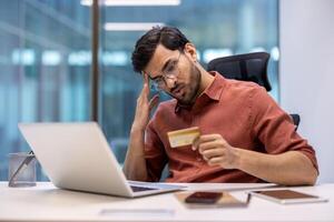 Man in office experiencing stress holding credit card while looking at laptop. Concept of financial issues, online transaction error, office work. Professional setting with modern workspace photo