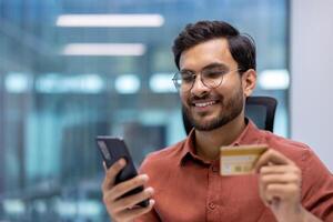 Business professional in modern office holding smartphone and credit card, smiling while making an online purchase. Represents digital payment, ecommerce, and technology in finance daily operations. photo