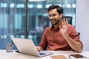 Friendly businessman waving during call using a laptop in modern office. Professional setting reflects positivity, communication, and productivity. Phone on table suggests digital connectivity. photo