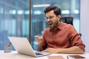 Business man showing frustration while pointing at laptop in office. Expresses emotions like anger or dismay during work. Seated at desk with stationery in modern environment. photo