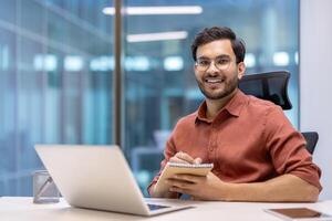 Smiling Hispanic businessman engaged in online meeting, using laptop, holding notebook in modern office. Demonstrates remote work and communication skills in professional setting. photo