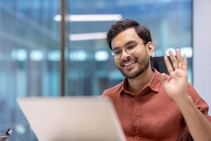 Latin American man businessman engaged in virtual meeting. Smiling while waving to colleague, sitting at office desk, using laptop for call. Modern communication, remote work concept. photo