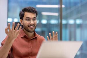 Young professional in modern office engaged in call, showing enthusiasm and gestures, wearing casual attire and eyeglasses, creating dynamic communication atmosphere. photo