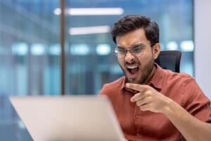 Surprised man with glasses pointing at laptop screen in office. Capturing shock and disbelief while working on computer. Office ambiance enhances feeling of unexpected discovery. photo