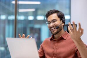Latin American man businessman in online meeting using laptop and headset at office desk. Expressing enthusiasm during remote work call, technology connection, communication, engagement. photo