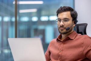 Young professional wearing headset working on laptop in modern office setting. Focused and engaged in online communication, displaying confidence. Formal attire suggests business environment. photo