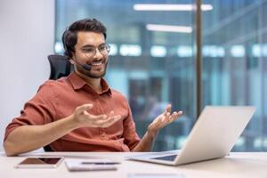 Businessman with headset in modern office conducting virtual meeting, smiling while communicating. Laptop open on desk, illustrating concept of tech, communication, success, business. photo