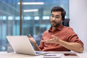 Young man wearing headphones engaged in call, using laptop, gesturing talking. Seated in modern office environment, digital devices on desk, conveying communication and technology concepts. photo