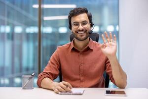 Hispanic businessman wearing headset sits in modern office with notepad and tablet, waving during virtual meeting. Shows professional, friendly communication skills and digital collaboration. photo