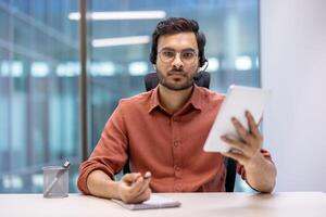 Latin American man businessman in office using digital tablet and headset for call. Engaged in remote communication, showcasing modern corporate technology and connectivity. photo