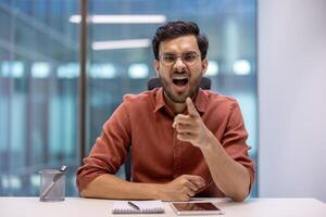 Hispanic businessman expressing frustration at desk, pointing assertively. Modern office setting with open background. Emotion conveyed through gestures, creating dynamic and intense atmosphere. photo