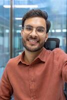 Young man with beard and glasses engaged in call in office. He is smiling warmly while looking directly at camera, capturing image with phone. Professional attire suggests office context. photo