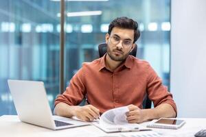 Confident businessman in modern office setting at desk with laptop, documents, phone. Focused expression suggests commitment, professionalism. Ideal for themes of work, productivity photo