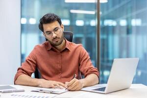 Young man in thoughtful expression working on important paperwork at desk. Displays focus and concentration. Modern office environment includes laptop and notepad for efficiency. photo
