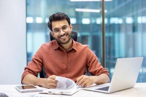 Businessman in an office environment smiling confidently while looking at documents. Laptop and phone visible, suggesting multitasking and productivity. Modern workspace background photo