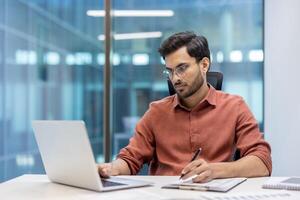 Confident man wearing glasses concentrating on laptop while taking notes. Engaged in serious paperwork, using digital tools for efficient work. Modern office environment enhances productivity focus. photo