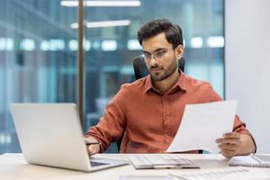 Confident man in glasses working with laptop and holding documents in office. Focused on screen and paperwork, demonstrating concentration and professionalism in business environment. photo