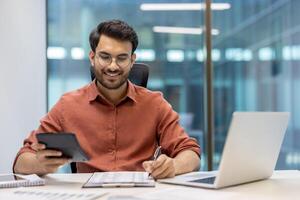 Smiling financier accountant reviews financial data with calculator and laptop. Professional efficiency displayed as he confidently tackles calculations and reports in modern office setting. Man exudes confidence and focus. photo